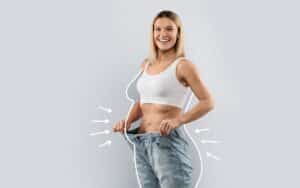 Woman in a sports bra holding out the waistband of loose jeans, with illustrated arrows and outline indicating weight loss and body contouring results