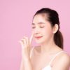 Beautiful Young Asian woman with clean fresh white skin touching her own nose softly with fingers in beauty pose. Girl closing her eyes, pink background. Facial treatment, cosmetology, surgery concept