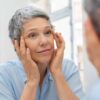 Choosing Between an Eyelid Lift and a Brow Lift Older woman looking at eyelids and forehead in mirror